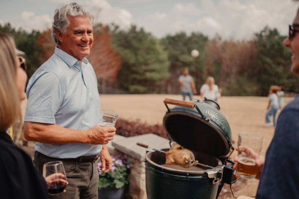 Man holding a drink standing next to a grill with Rotisserie on it, outdoors.