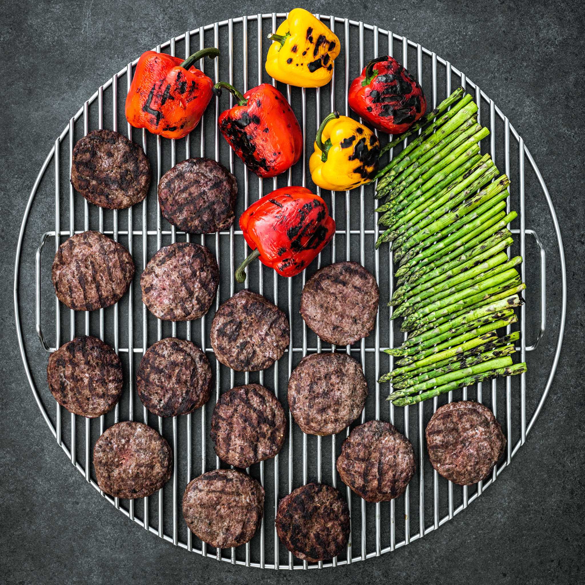 Overhead view of a 2XL Big Green Egg cooking grid loaded edge to edge with burgers, charred peppers and asparagus
