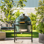 Large green ceramic kamado grill on a stand outdoors with trees and a building in the background
