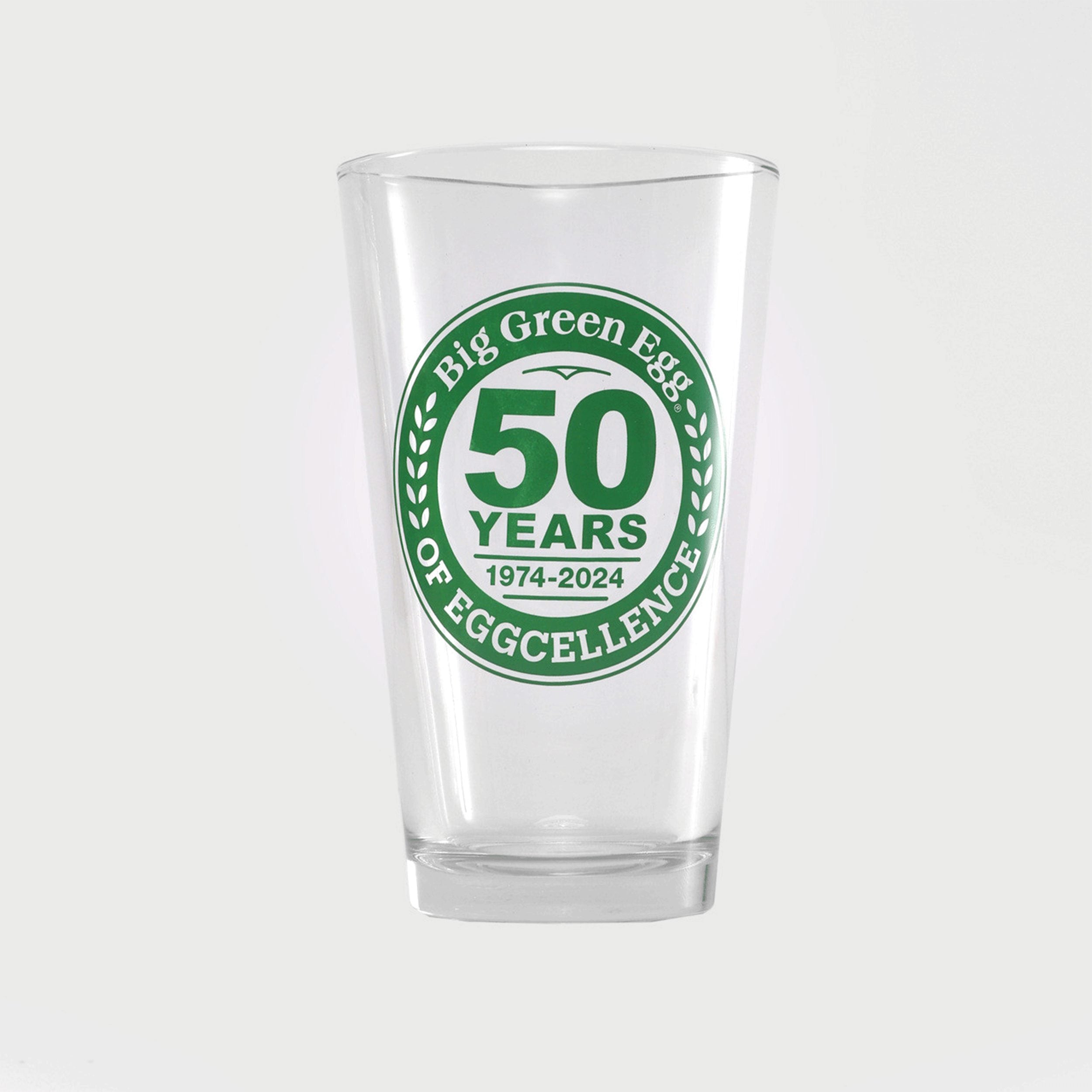 A clear drinking glass with a green circular 'Big Green Egg' logo featuring '50 YEARS OF EGGCELLENCE' and '1974-2024' printed on the front, set against a light gray background.