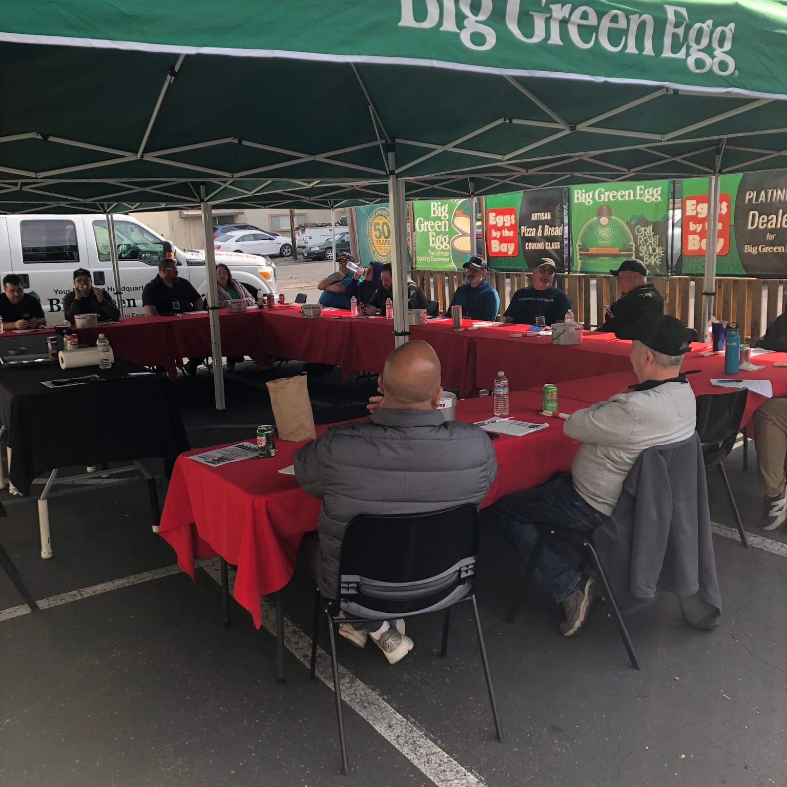 People sitting at tables under a green 'Big Green Egg' umbrella in an outdoor setting.