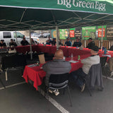 People sitting at tables under a green 'Big Green Egg' umbrella in an outdoor setting.