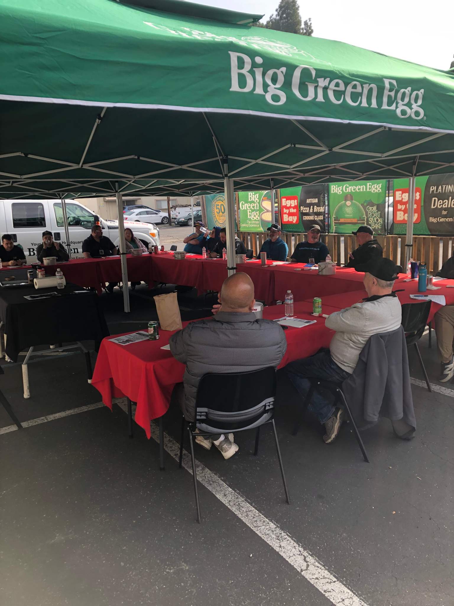 People sitting at tables under a green 'Big Green Egg' umbrella in an outdoor setting.