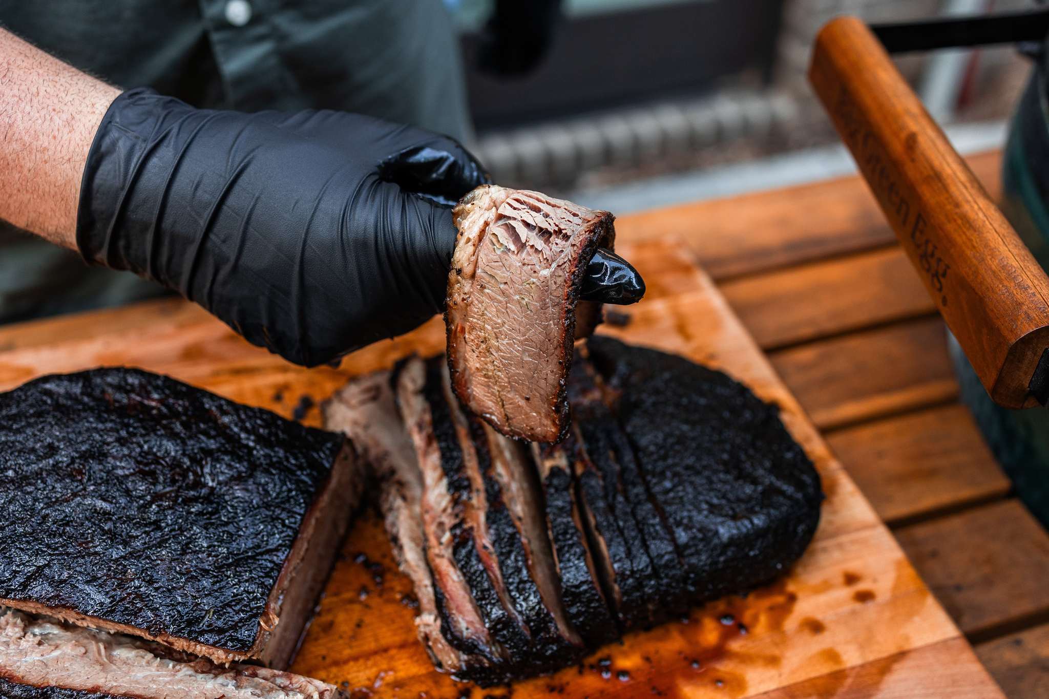 Hand wearing a black glove holding a piece of smoked meat on a wooden cutting board.