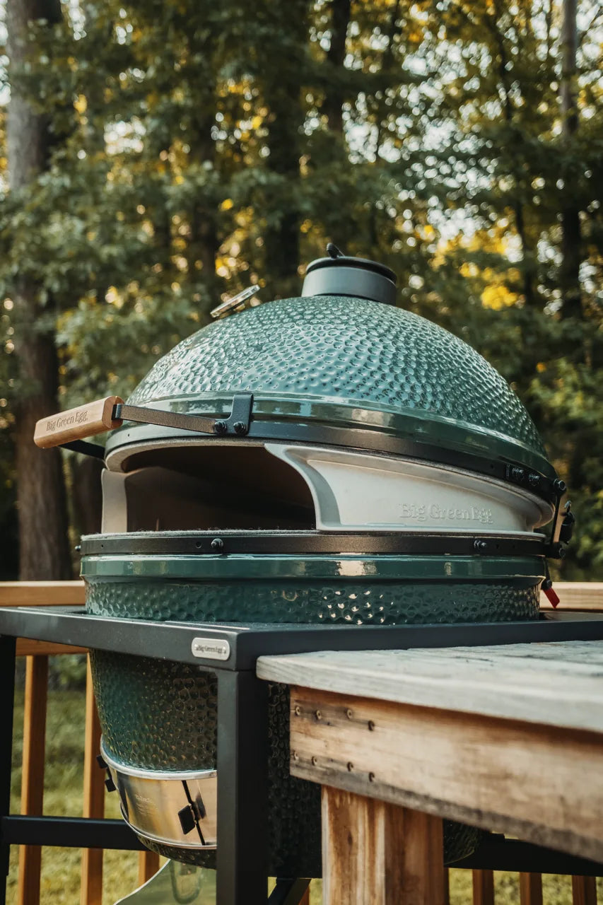 Green outdoor grill functioning as a pizza oven on a wooden table with trees in the background