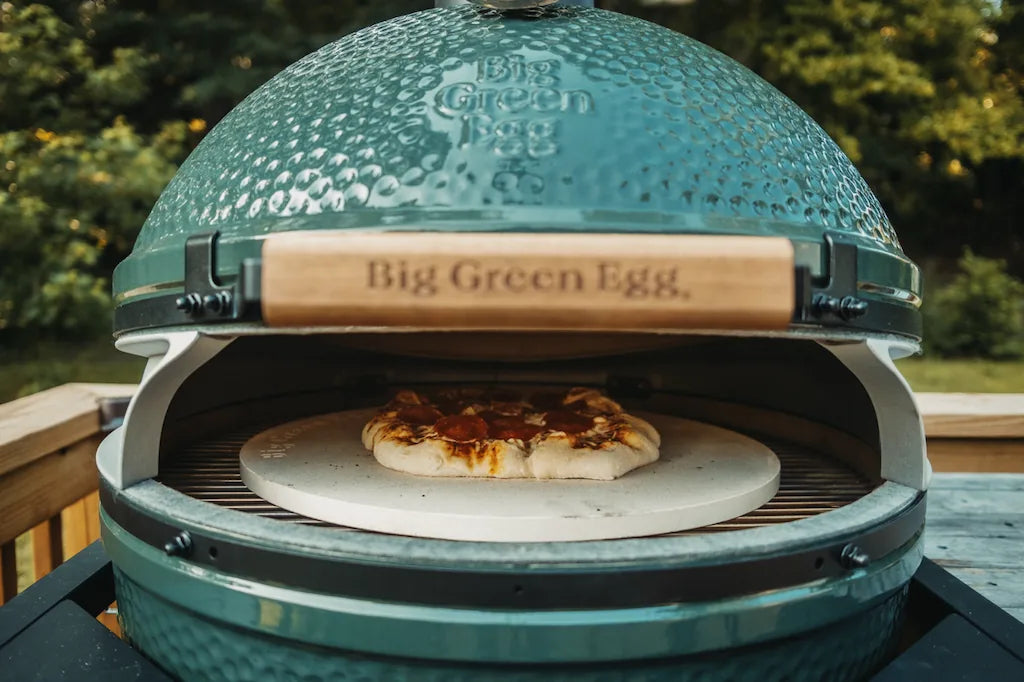 Big Green Egg ceramic kamado grill with a pizza inside, set against a natural outdoor background.