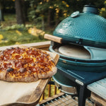 Wood-fired pizza on a wooden board with a large green ceramic pizza oven grill in the background.