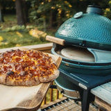Wood-fired pizza on a wooden board with a large green ceramic pizza oven grill in the background.