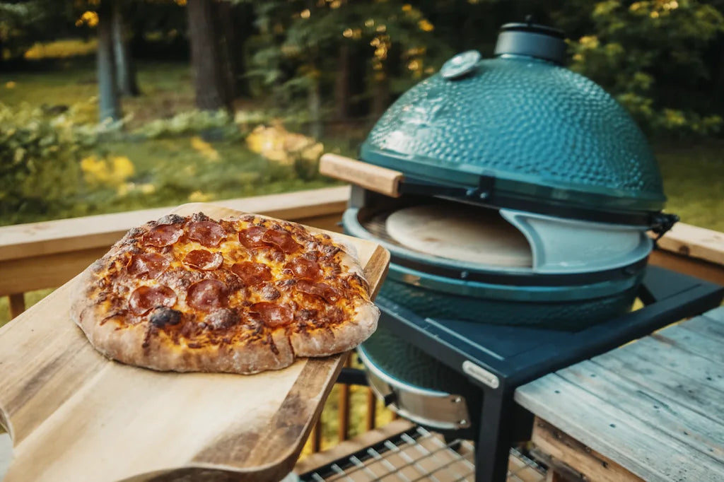 Wood-fired pizza on a wooden board with a large green ceramic pizza oven grill in the background.