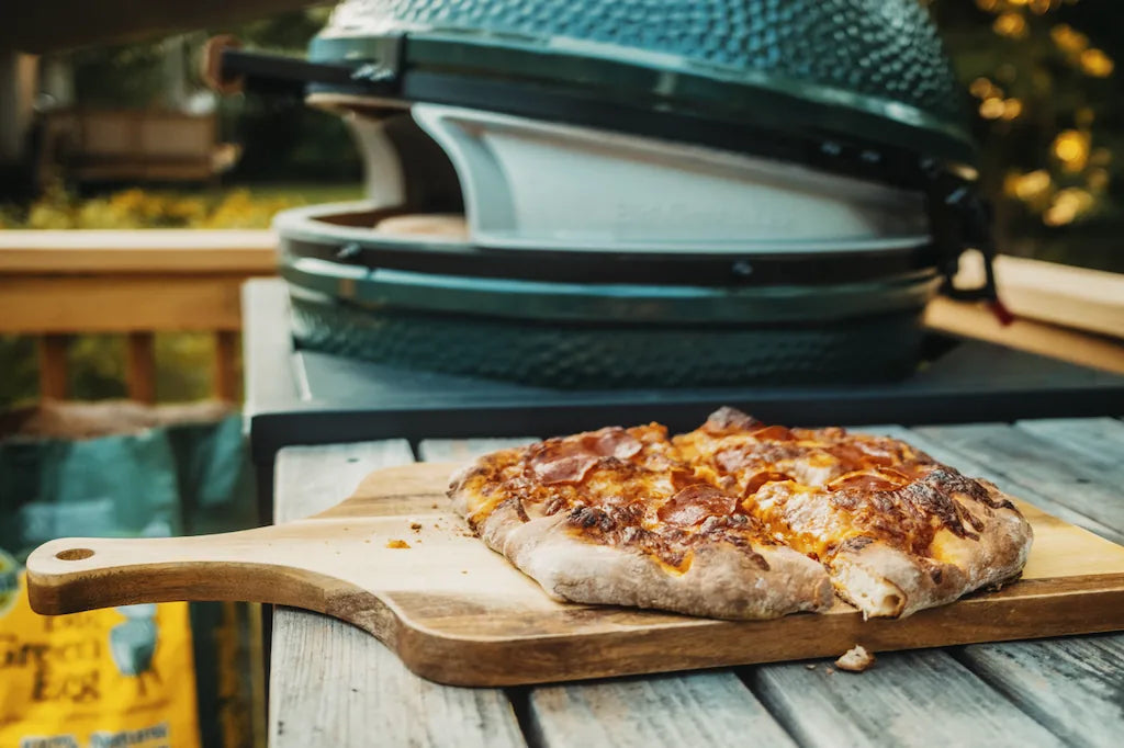 Wood-fired pizza on a wooden cutting board in front of a large green barbecue grill.