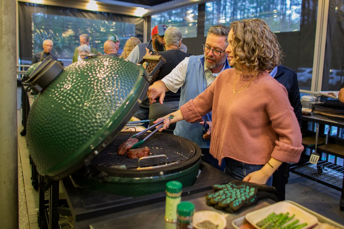 People grilling food on a large green ceramic barbecue grill in an indoor setting.