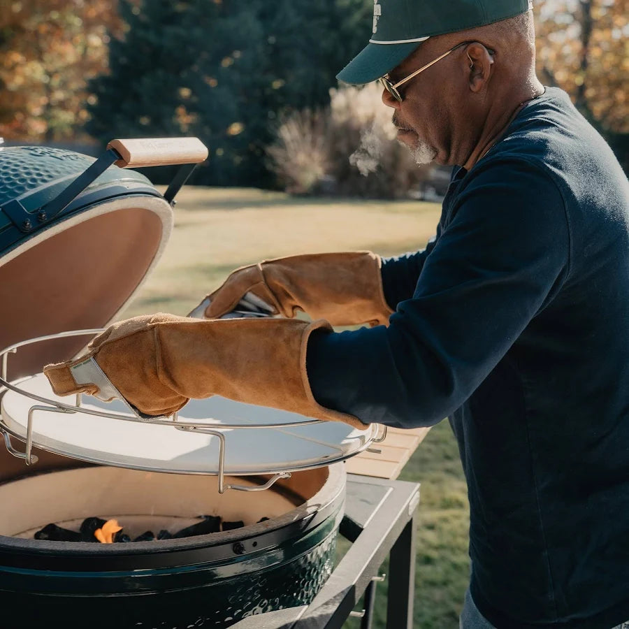 A man wearing a green cap, sunglasses, a navy long-sleeve shirt, and tan leather grill gloves is tending a ceramic kamado grill outdoors in a sunny backyard.