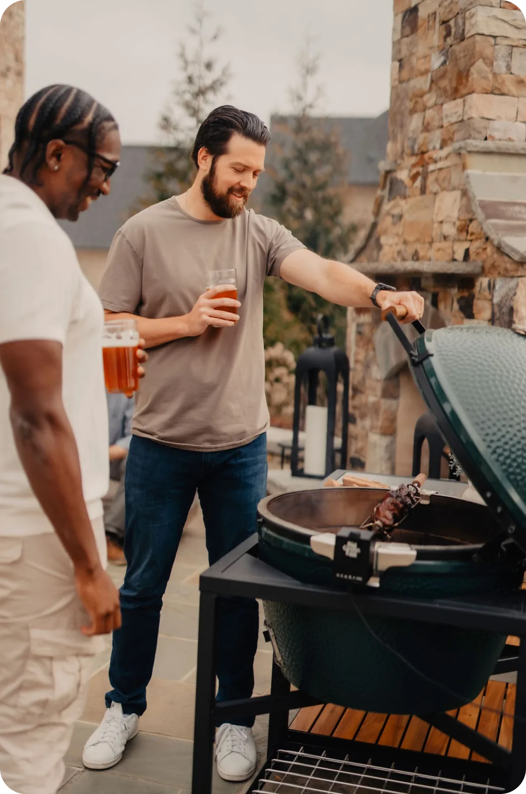 Two men standing outdoors by a grill, one holding a beer.