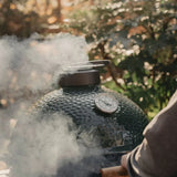 Smoking Big Green Egg ceramic kamado barbecue grill outdoors with trees in the background
