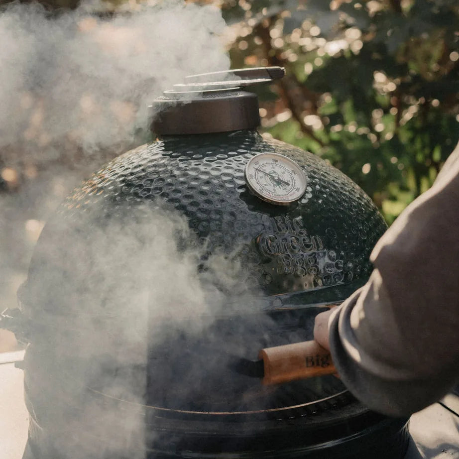 Smoking Big Green Egg ceramic kamado grill with a visible brand logo, outdoors and surrounded by greenery