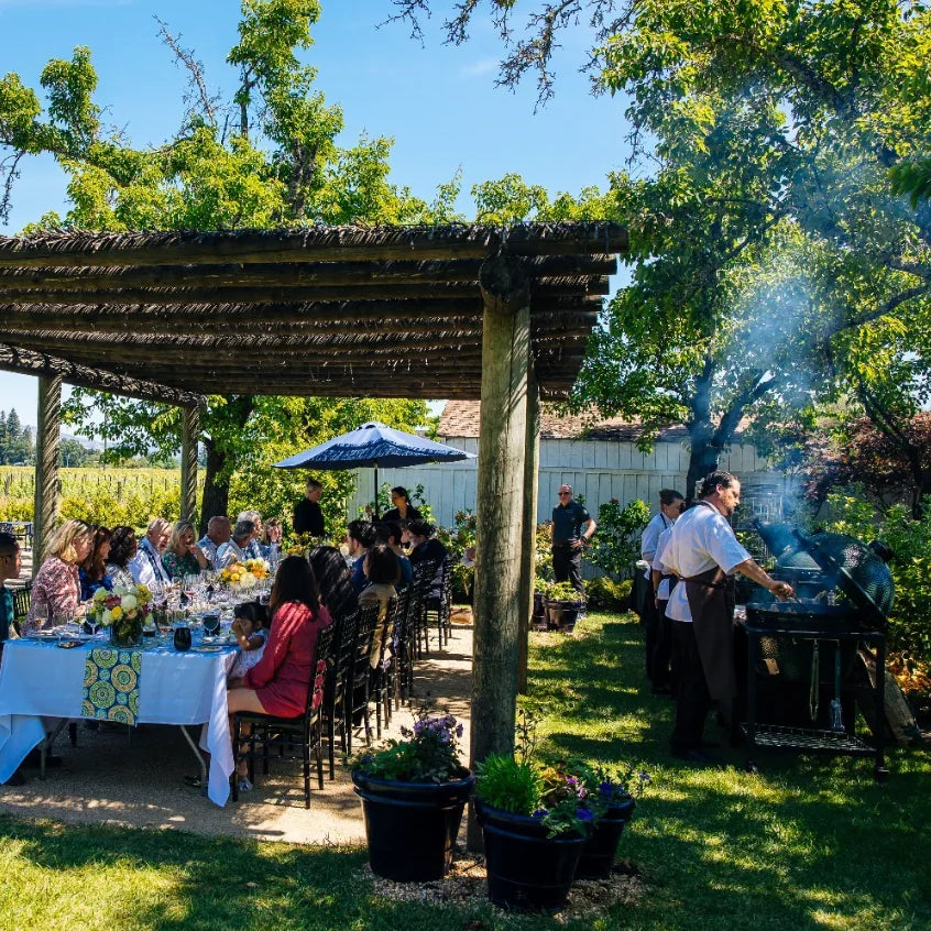 Outdoor gathering with people seated at tables under a wooden pergola, kamado grill in the background
