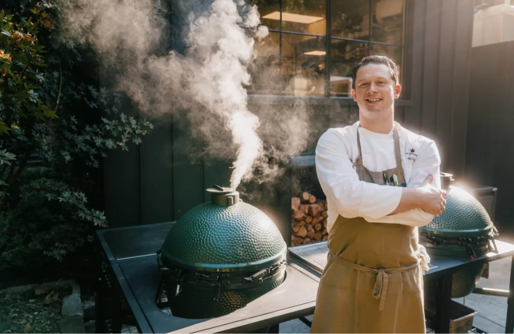 Man in an apron standing next to a large green ceramic kamado barbecue grill with smoke rising