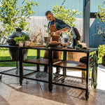 Man preparing food outdoors at a large black outdoor kitchen table with a grill and various items.