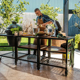 Man preparing food outdoors at a large black outdoor kitchen table with a grill and various items.