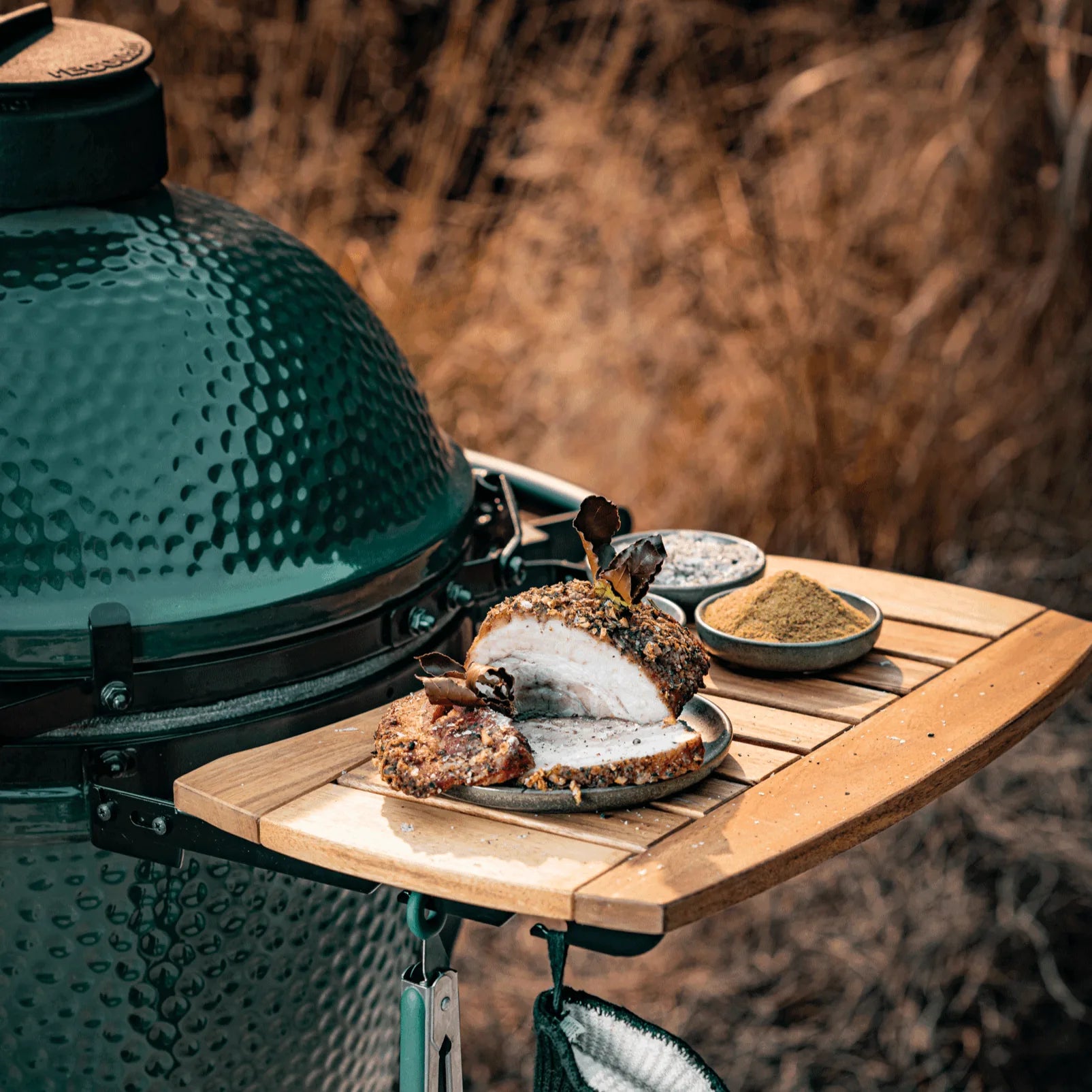 Big Green Egg ceramic kamado grill with a wooden cutting board displaying sliced meat and herbs outdoors.