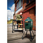 Person preparing food next to a large green ceramic kamado barbecue grill on a wooden deck.