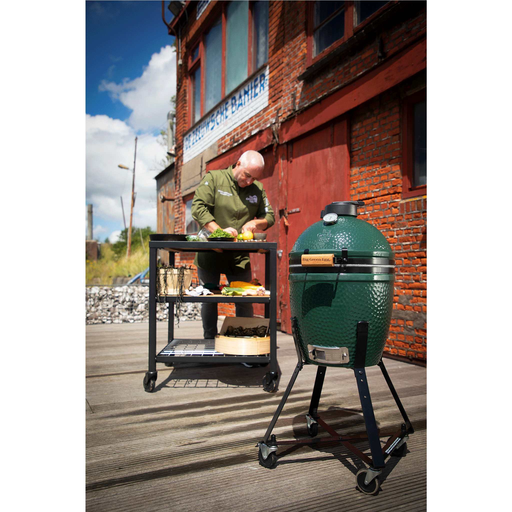 Person preparing food next to a large green ceramic kamado barbecue grill on a wooden deck.