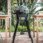 Green Egg ceramic kamado barbecue grill on a stand outdoors with tables and plants in the background