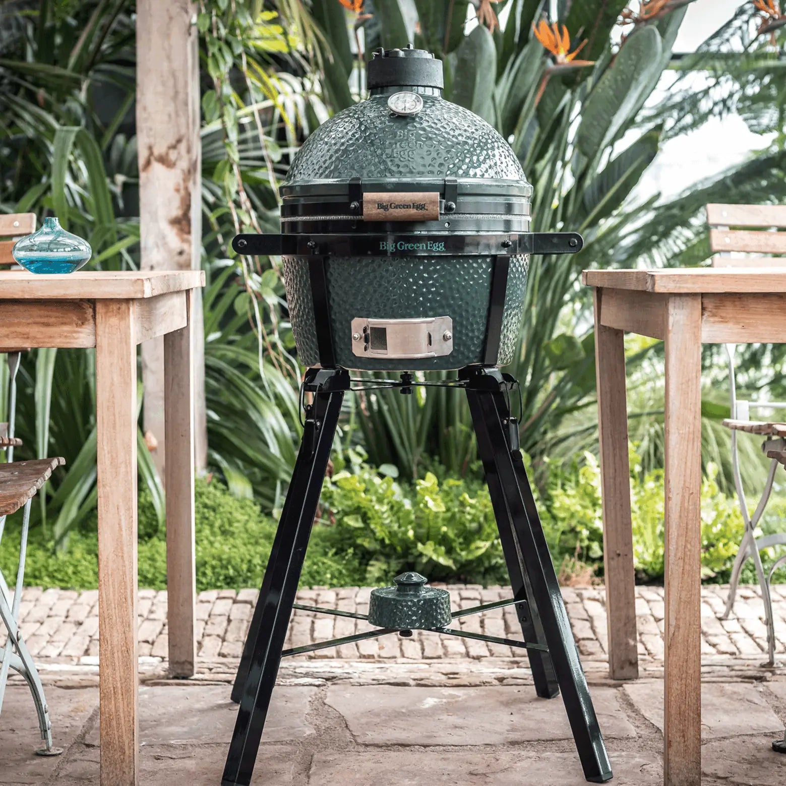 Green Egg ceramic kamado barbecue grill on a stand outdoors with tables and plants in the background