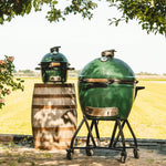 Two green Kamado-style ceramic grills outdoors with a wooden barrel and field background.
