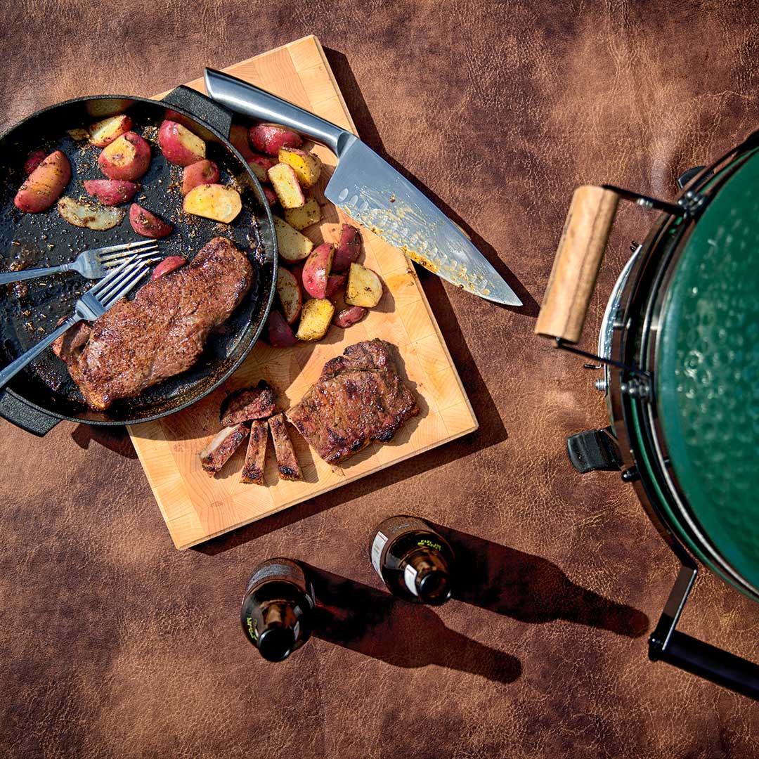 Cast Iron Skillet with steak, potatoes, forks and knife on cutting and Big Green Egg.